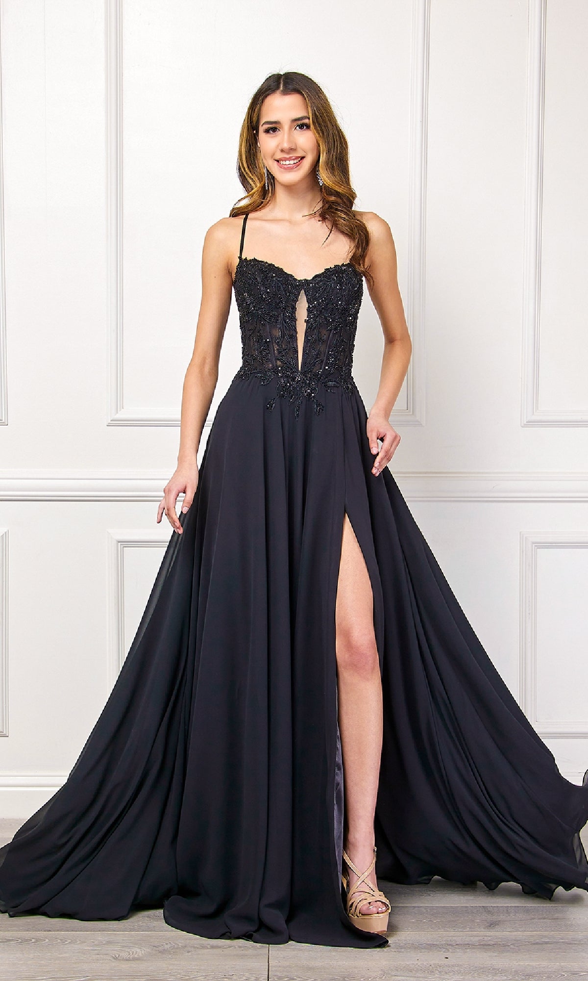 A woman in the Amelia Couture 7066 Long Black Corset Prom Dress, featuring a lace bodice, spaghetti straps, front cutout, and thigh-high slit, stands indoors against a white paneled wall.